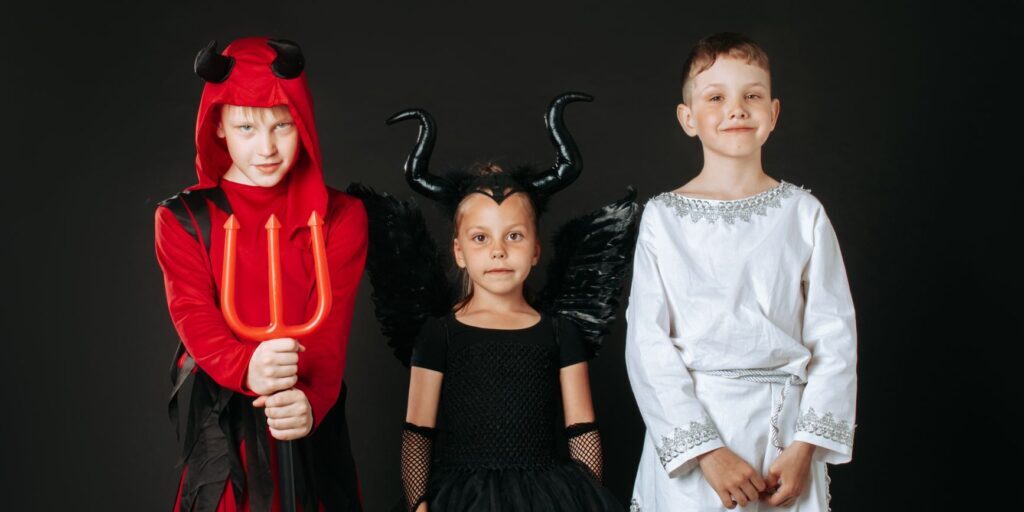 Three children wear Halloween costumes: devil, angel, and maleficent on a dark background.